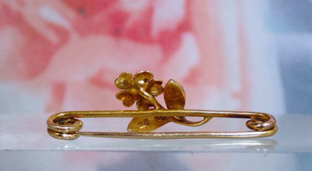 A Victorian 15ct gold bar brooch featuring a central textured flower set with a diamond and foliage set with seed pearls. View V