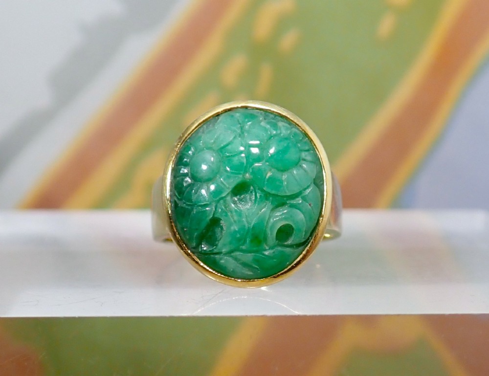 A mid-century yellow gold ring featuring a smooth bezel setting holding an oval carved green jadeite gemstone with floral detail.
