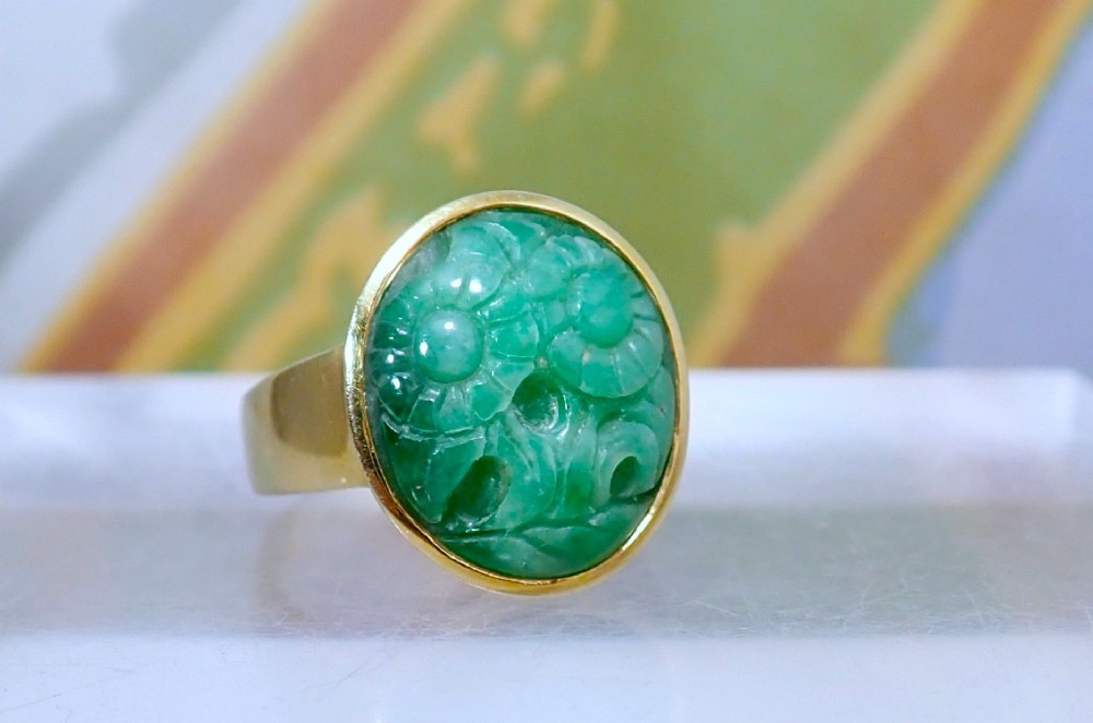 A mid-century yellow gold ring featuring a smooth bezel setting holding an oval carved green jadeite gemstone with floral detail. View II