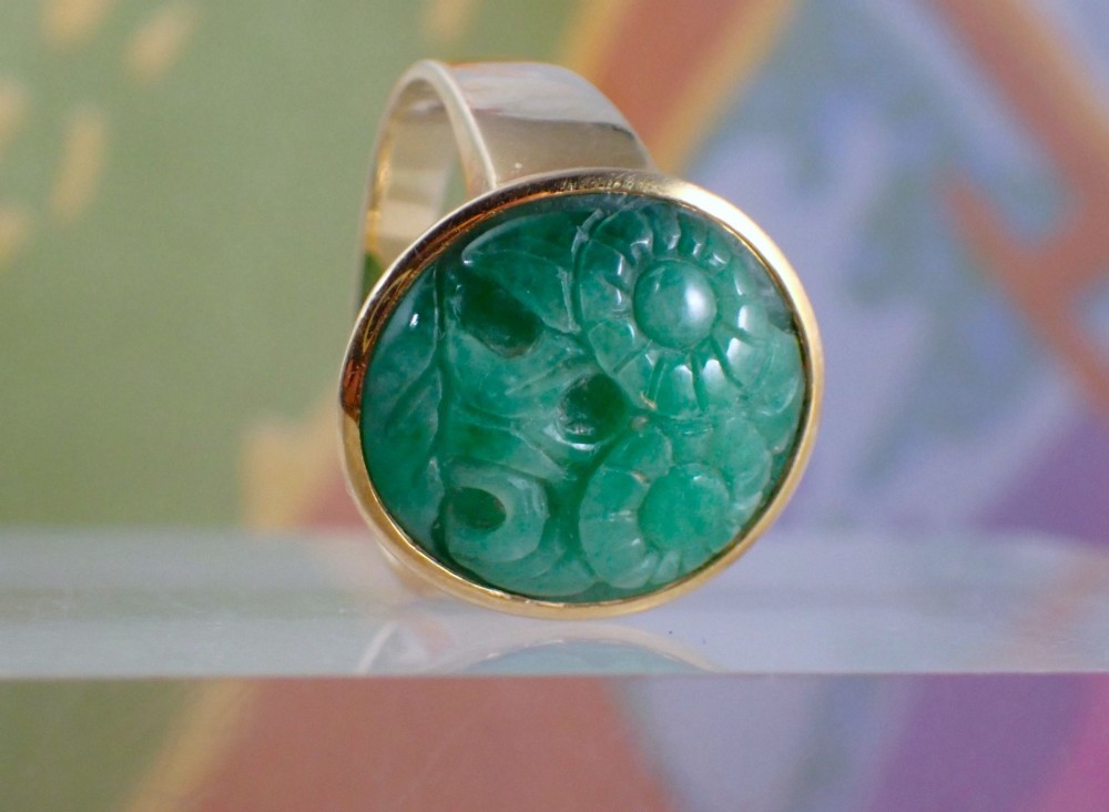 A mid-century yellow gold ring featuring a smooth bezel setting holding an oval carved green jadeite gemstone with floral detail. View V