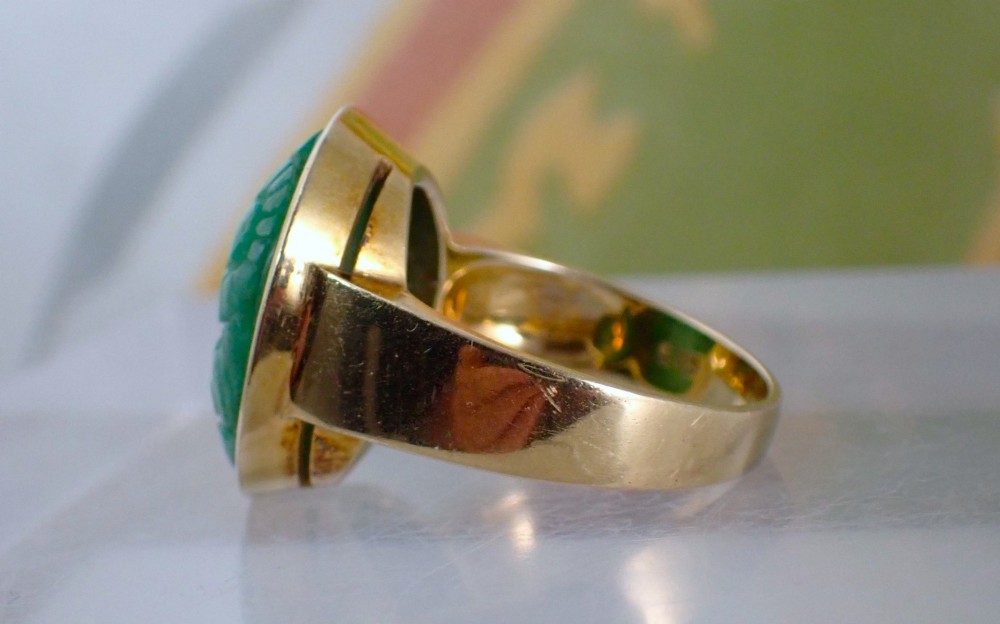 A mid-century yellow gold ring featuring a smooth bezel setting holding an oval carved green jadeite gemstone with floral detail. View VI
