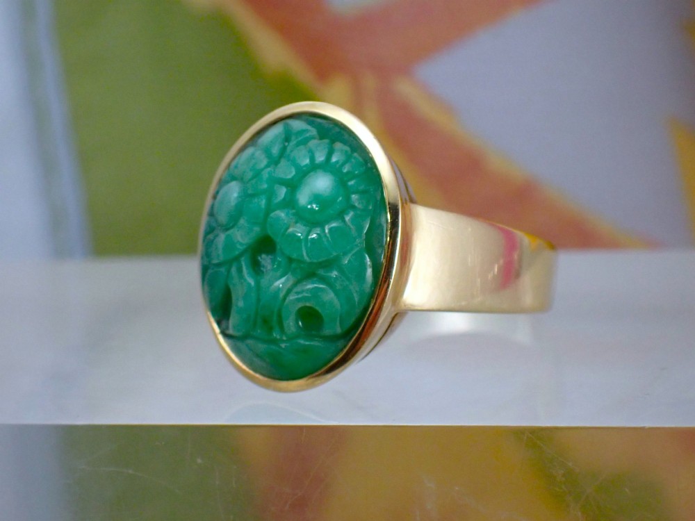 A mid-century yellow gold ring featuring a smooth bezel setting holding an oval carved green jadeite gemstone with floral detail. View IX