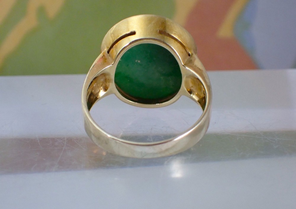A mid-century yellow gold ring featuring a smooth bezel setting holding an oval carved green jadeite gemstone with floral detail. View X