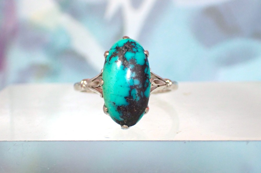 An Art Deco 18ct white gold ring featuring a central oval turquoise cabochon with natural dark matrix patterning, held by claw settings above decorative pierced shoulders.
