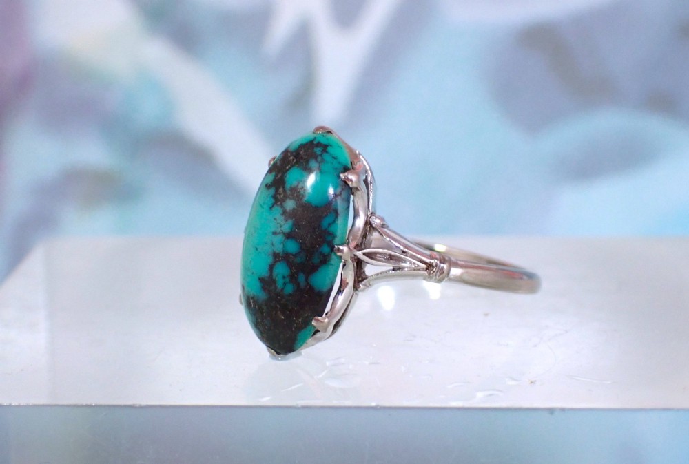 An Art Deco 18ct white gold ring featuring a central oval turquoise cabochon with natural dark matrix patterning, held by claw settings above decorative pierced shoulders. View II