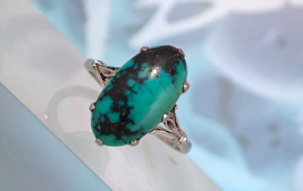 An Art Deco 18ct white gold ring featuring a central oval turquoise cabochon with natural dark matrix patterning, held by claw settings above decorative pierced shoulders. View VII
