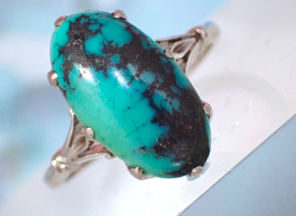 An Art Deco 18ct white gold ring featuring a central oval turquoise cabochon with natural dark matrix patterning, held by claw settings above decorative pierced shoulders. View VIII