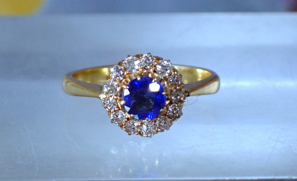 An Edwardian 18ct yellow gold ring featuring a central round blue sapphire surrounded by a floral halo of small diamonds. View II