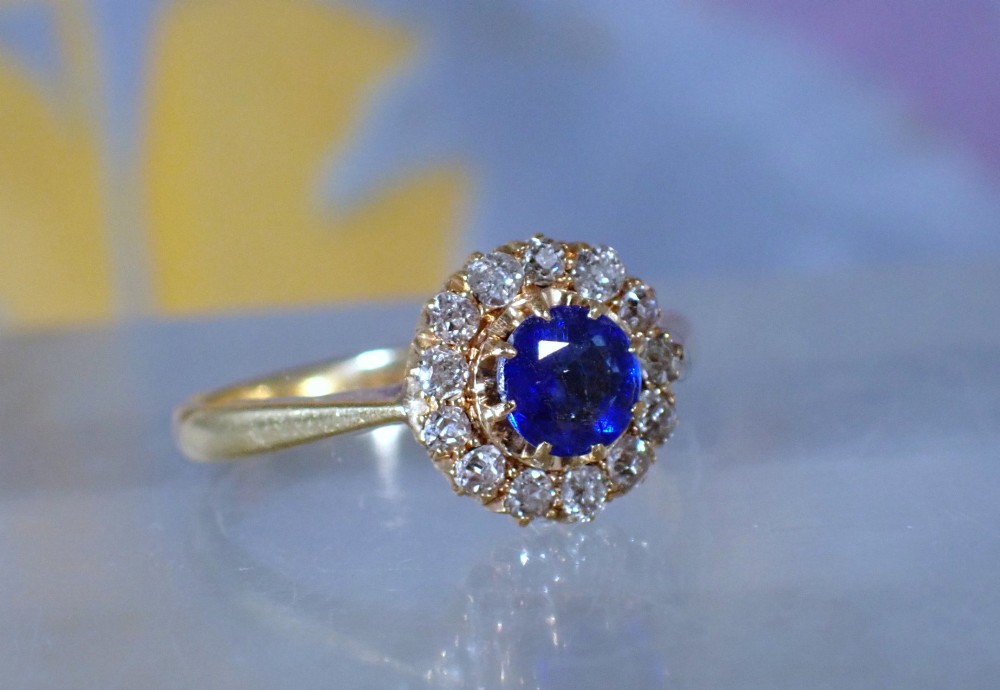 An Edwardian 18ct yellow gold ring featuring a central round blue sapphire surrounded by a floral halo of small diamonds. View III