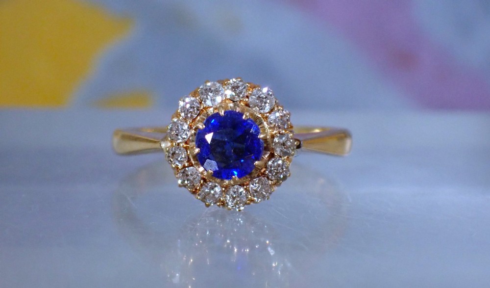 An Edwardian 18ct yellow gold ring featuring a central round blue sapphire surrounded by a floral halo of small diamonds. View IV