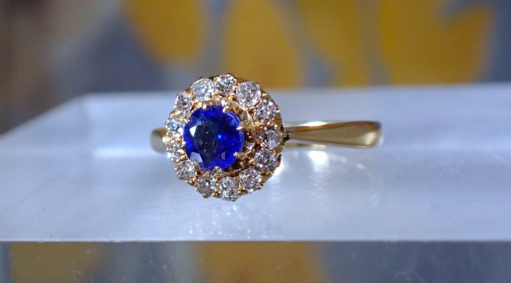 An Edwardian 18ct yellow gold ring featuring a central round blue sapphire surrounded by a floral halo of small diamonds. View V