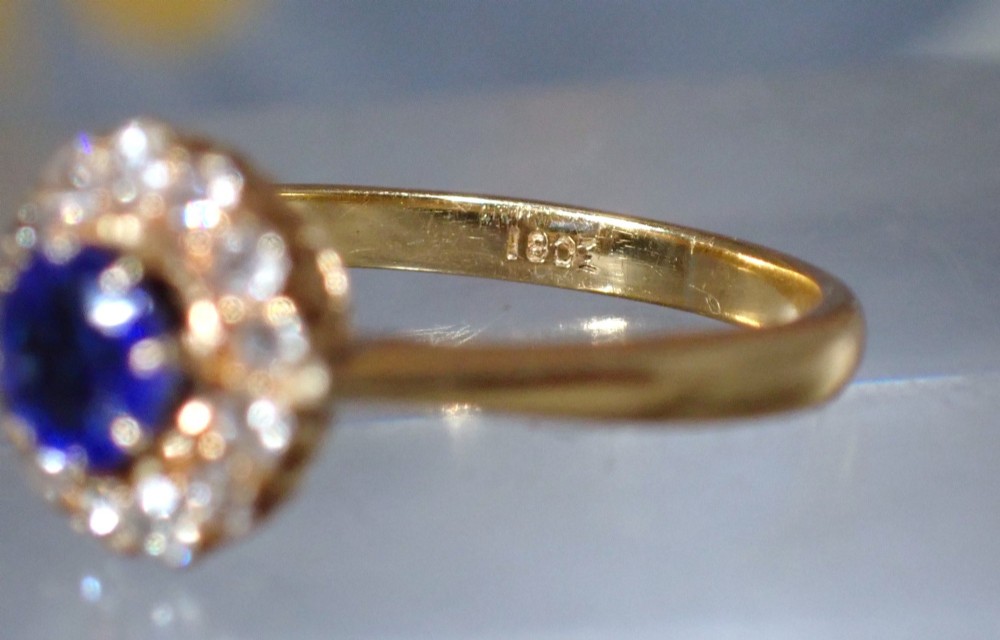 An Edwardian 18ct yellow gold ring featuring a central round blue sapphire surrounded by a floral halo of small diamonds. View VI