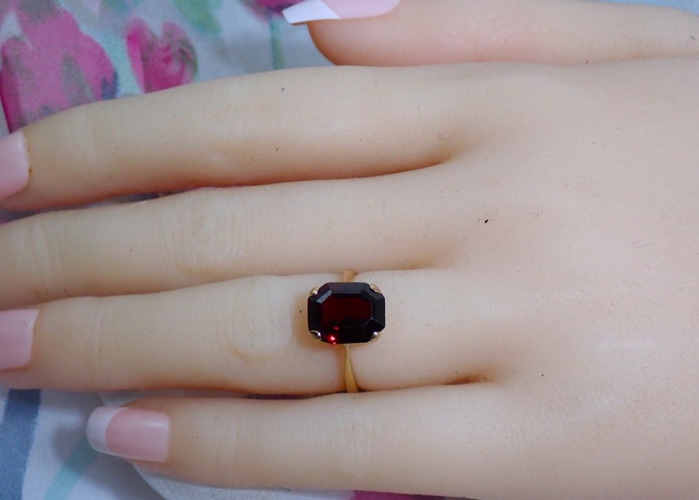 An Art Deco style 9ct yellow gold ring featuring a large, rectangular emerald-cut deep red garnet held by four corner prongs on a slender gold band. View VI