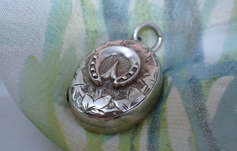 An oval sterling silver locket decorated with a central relief horseshoe motif surrounded by hand-engraved leaf patterns and fitted with a top jump ring. View II