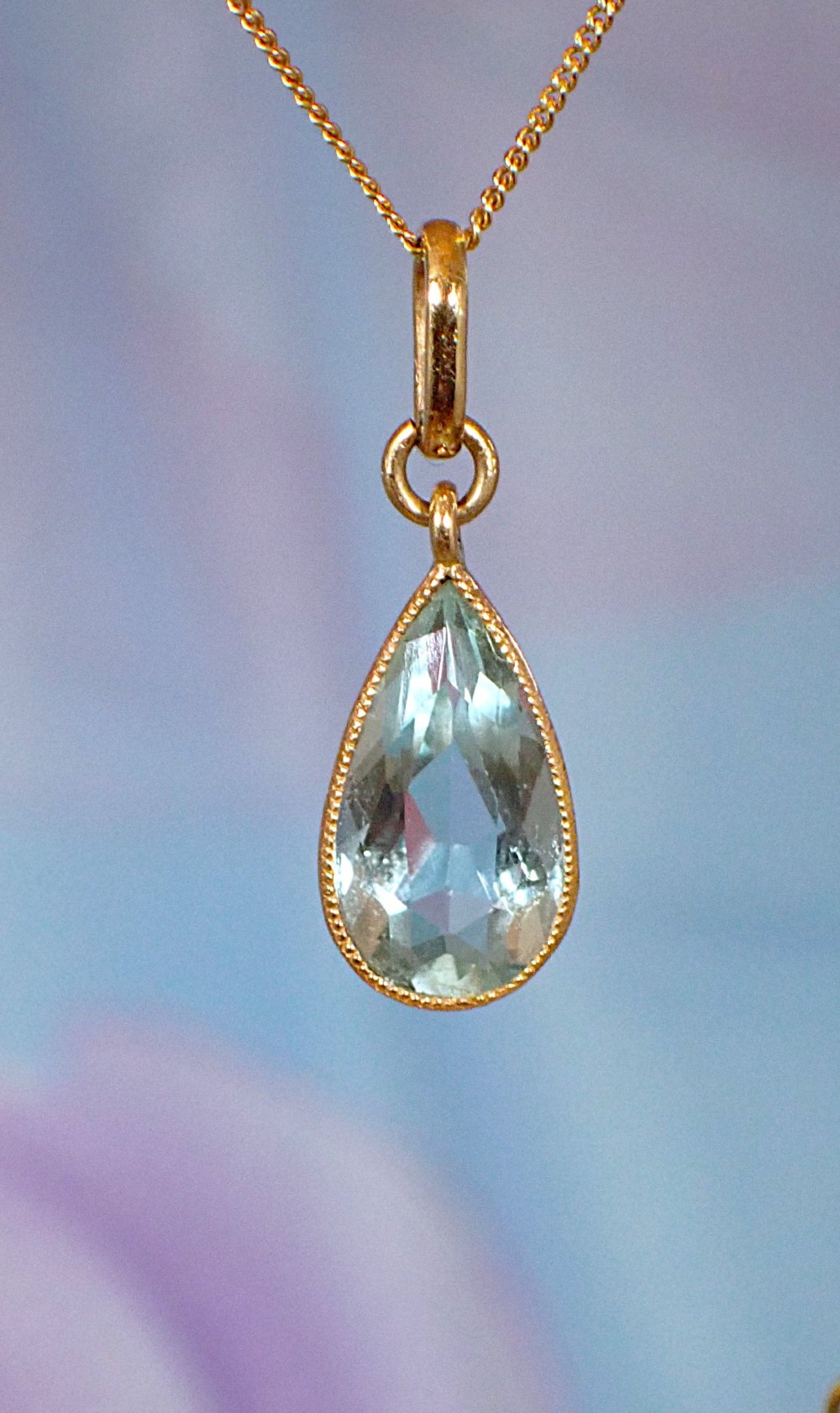 A pear-shaped pale blue aquamarine gemstone set in a yellow gold bezel pendant suspended from a fine gold chain.