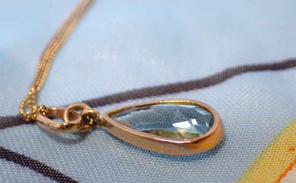 A pear-shaped pale blue aquamarine gemstone set in a yellow gold bezel pendant suspended from a fine gold chain. View IV
