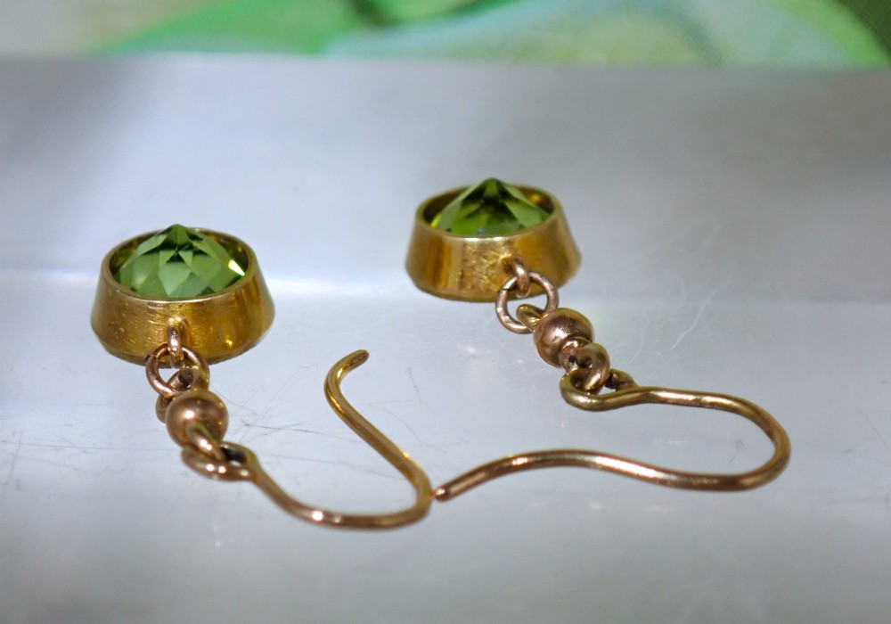 Pair of antique Edwardian 15ct gold drop earrings featuring circular faceted green peridots in collet settings suspended from wire hooks. View V