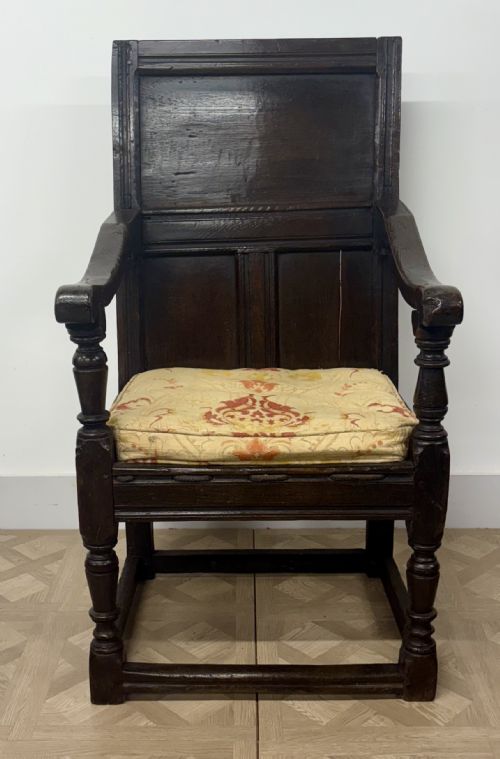 fig 2. A 17th-century dark oak wainscot armchair featuring a solid panelled back, turned front supports, sturdy stretchers, and a patterned fabric seat cushion.