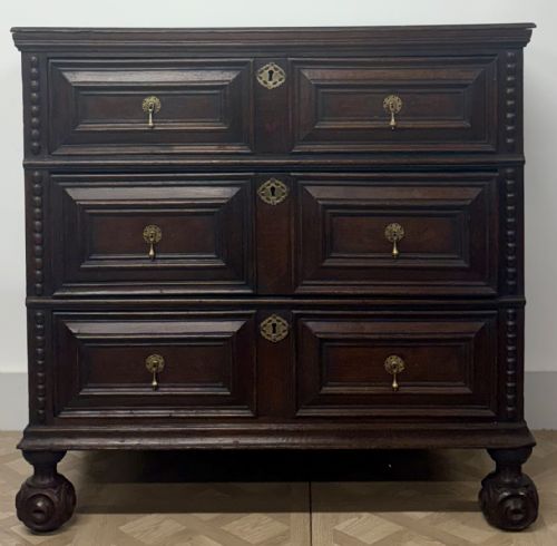 fig 1. A late seventeenth-century dark oak chest of drawers featuring three geometrically panelled drawer fronts, brass teardrop handles, central escutcheons, and turned bun feet.
