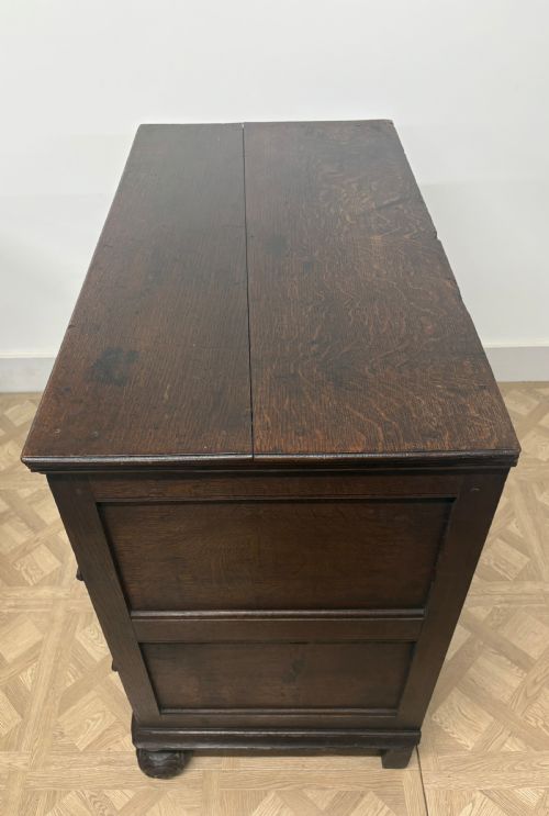 fig 3. A late seventeenth-century dark oak chest of drawers featuring three geometrically panelled drawer fronts, brass teardrop handles, central escutcheons, and turned bun feet.