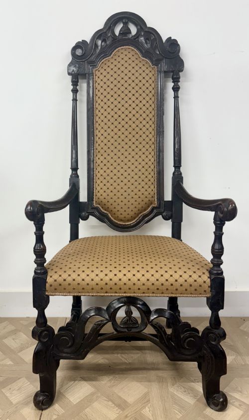 fig 1. A late 17th-century Carolean carved dark walnut armchair featuring a tall padded backrest, scrolling arms, gold patterned upholstery, and an ornate carved stretcher.