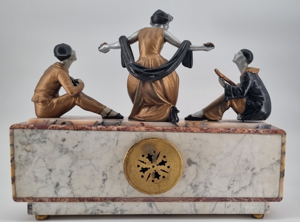An Art Deco mantel clock featuring a veined marble base with a rectangular brass dial, surmounted by three patinated metallic figures depicting musicians and a dancer. View VIII