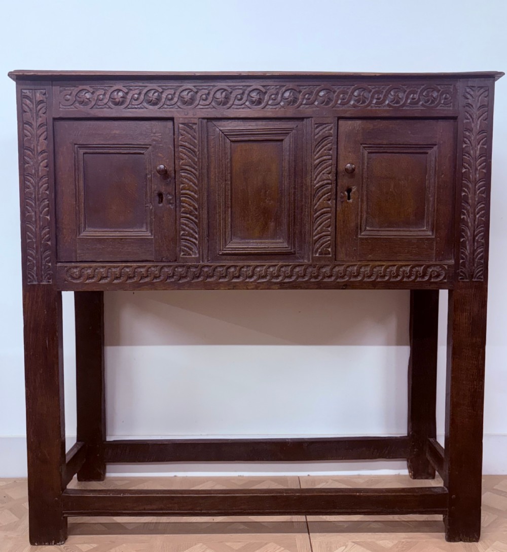 A 17th-century joined dark oak cupboard on stand featuring carved guilloche friezes, panelled doors, and square stile legs joined by stretchers. View II