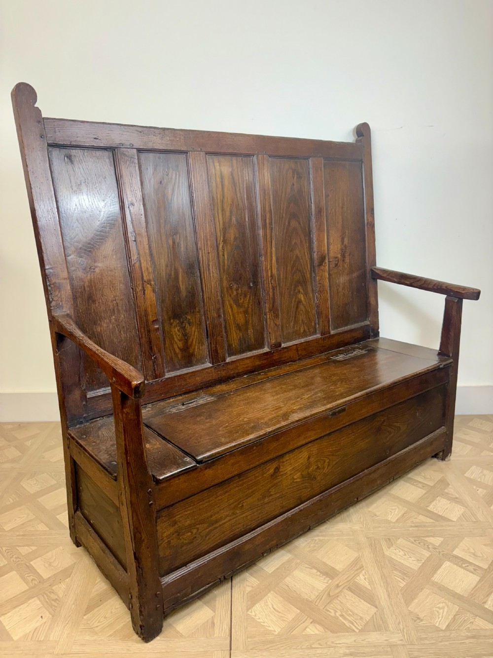 An 18th-century oak and ash box settle featuring a high five-panel back, straight armrests, and a hinged bench seat over a solid storage base. View V