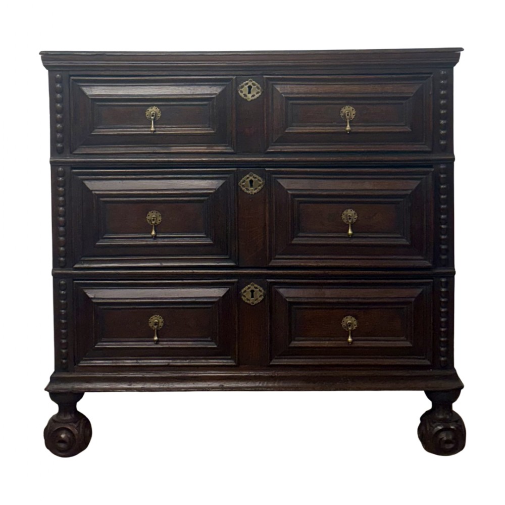 A late seventeenth-century dark oak chest of drawers featuring three geometrically panelled drawer fronts, brass teardrop handles, central escutcheons, and turned bun feet.