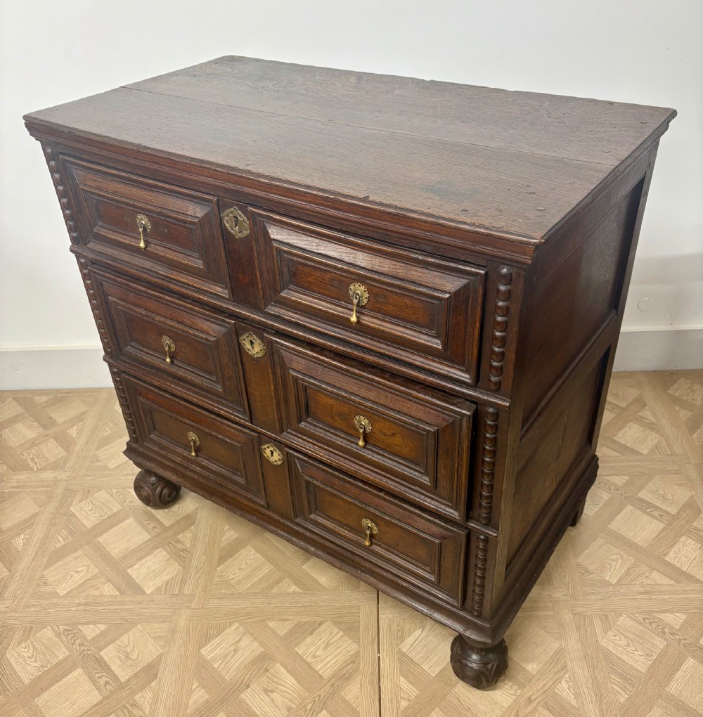 A late seventeenth-century dark oak chest of drawers featuring three geometrically panelled drawer fronts, brass teardrop handles, central escutcheons, and turned bun feet. View III