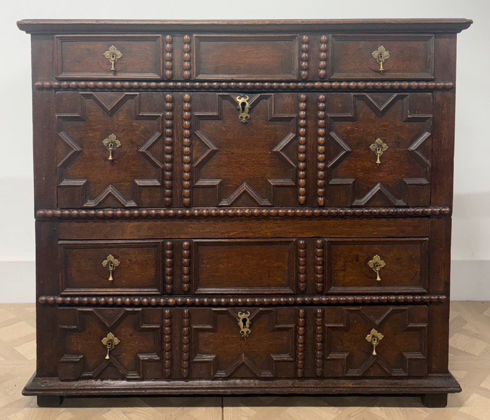A seventeenth-century dark oak chest of drawers featuring geometric applied mouldings, bobbin detailing, and brass drop handles. View VI