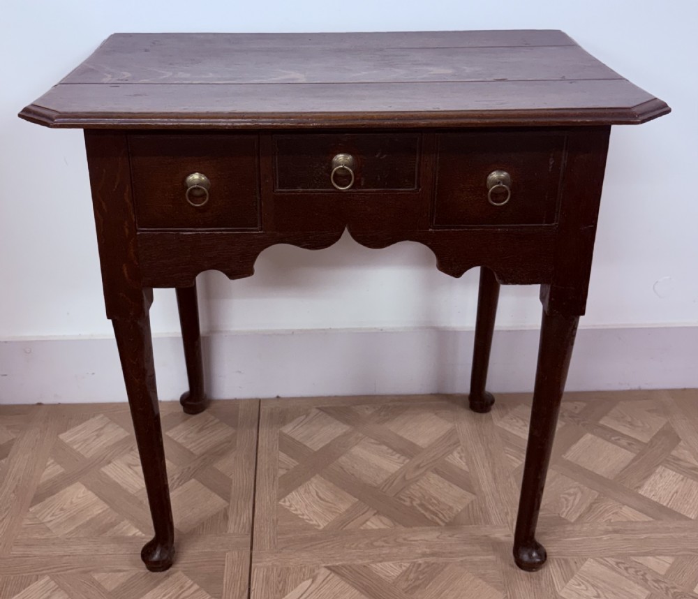 An early 18th-century dark oak side table featuring three drawers with brass ring pulls, a scalloped apron, and slender cabriole legs ending in pad feet.