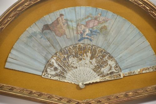 fig 1. A nineteenth-century hand-painted fan depicting classical figures, mounted inside a fan-shaped gilt wood frame against an ochre background.