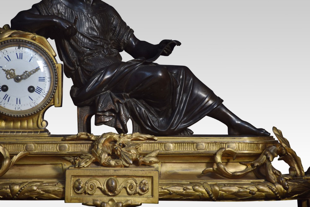 A late 19th-century French gilt metal mantel clock featuring a dark bronze classical figure seated beside an enamel dial on an ornate gilded scroll base. View VI