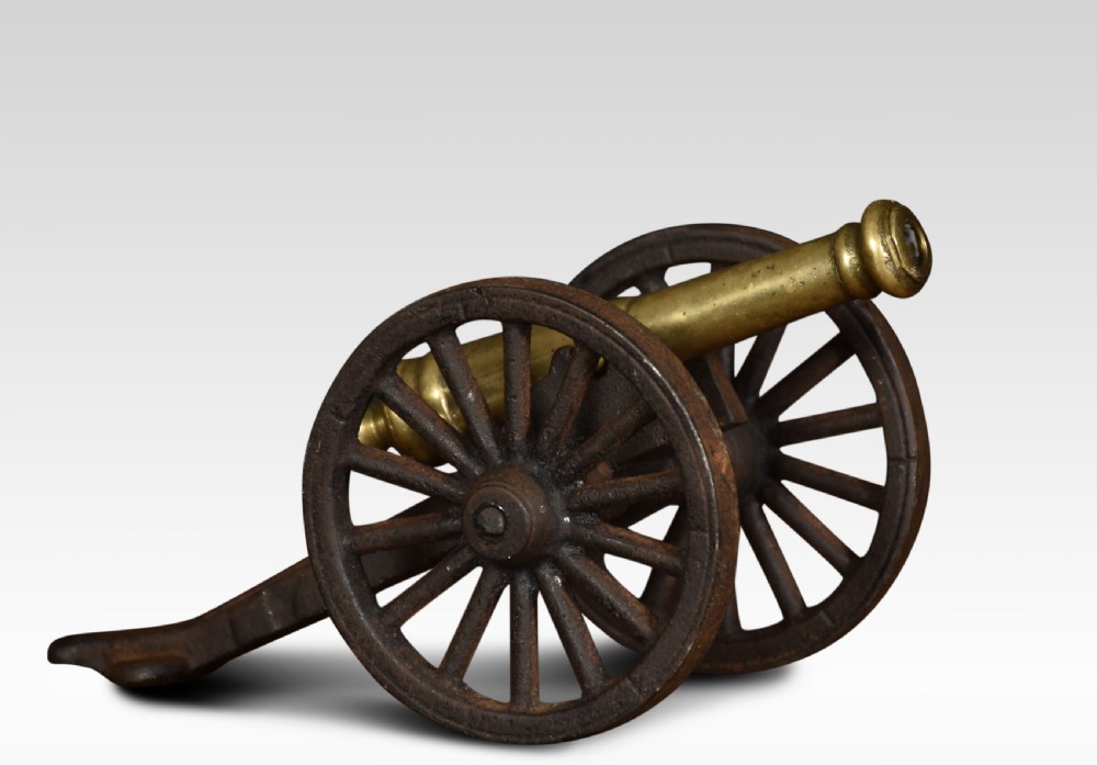 A miniature brass signal cannon barrel mounted on a dark cast iron two-wheeled carriage.