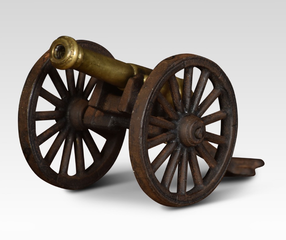 A miniature brass signal cannon barrel mounted on a dark cast iron two-wheeled carriage. View II