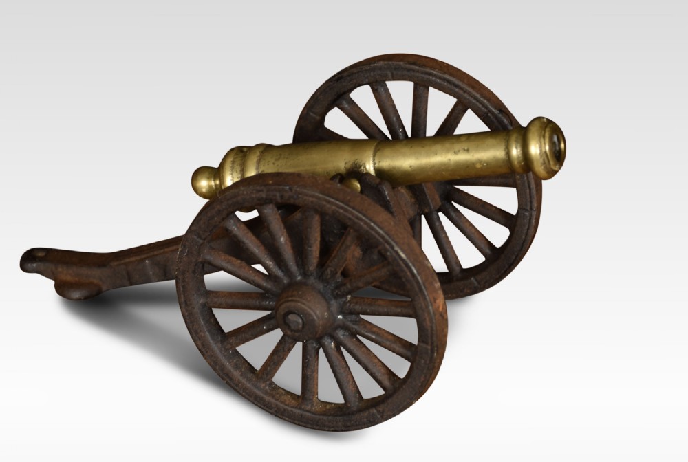 A miniature brass signal cannon barrel mounted on a dark cast iron two-wheeled carriage. View IV