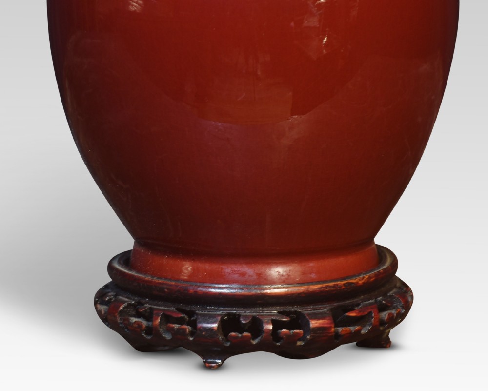 An oxblood red Chinese porcelain baluster vase fitted as a table lamp base, resting on a dark carved wooden plinth with brass double socket fittings. View II