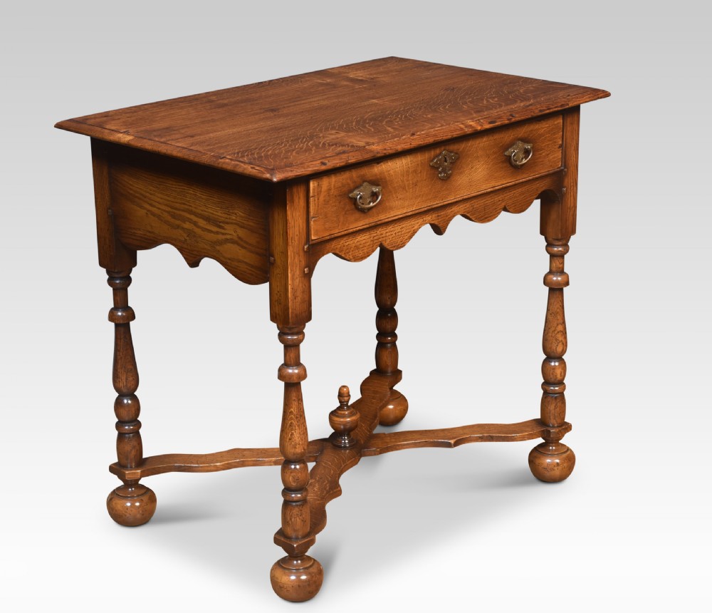 A dark oak side table featuring a single drawer with brass handles, a scalloped apron, turned legs, and an X-shaped cross stretcher.