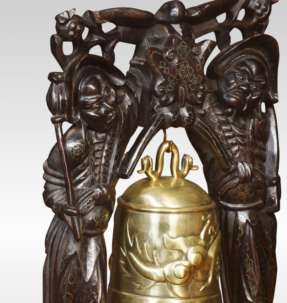A metallic brass bell suspended within an arch carved from dark hardwood featuring two standing figures on a molded wooden base. View II