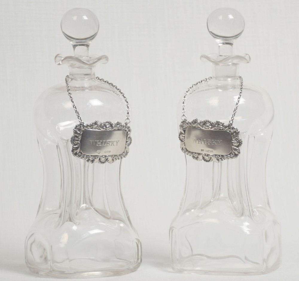 A pair of clear glass decanters featuring spherical stoppers, flared rims, and silver-coloured labels engraved with the word WHISKY suspended from fine chains.