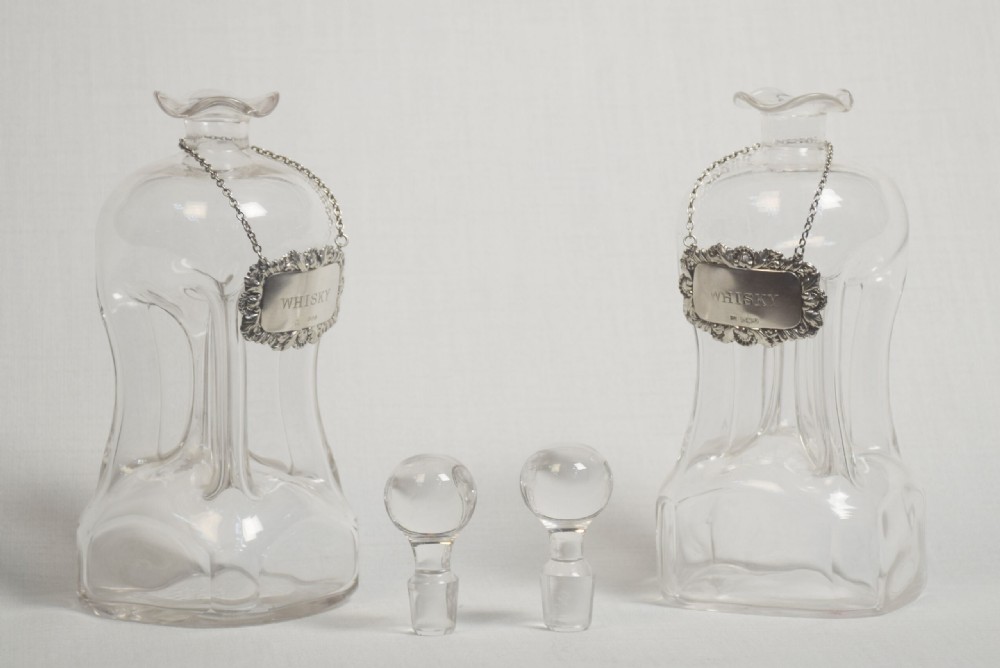 A pair of clear glass decanters featuring spherical stoppers, flared rims, and silver-coloured labels engraved with the word WHISKY suspended from fine chains. View IV