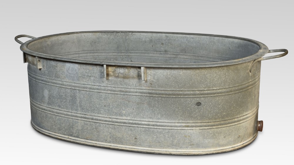 An oval galvanised steel bath tub featuring side carrying handles, ribbed horizontal reinforcement bands, a rolled rim, and a drainage plug near the base.