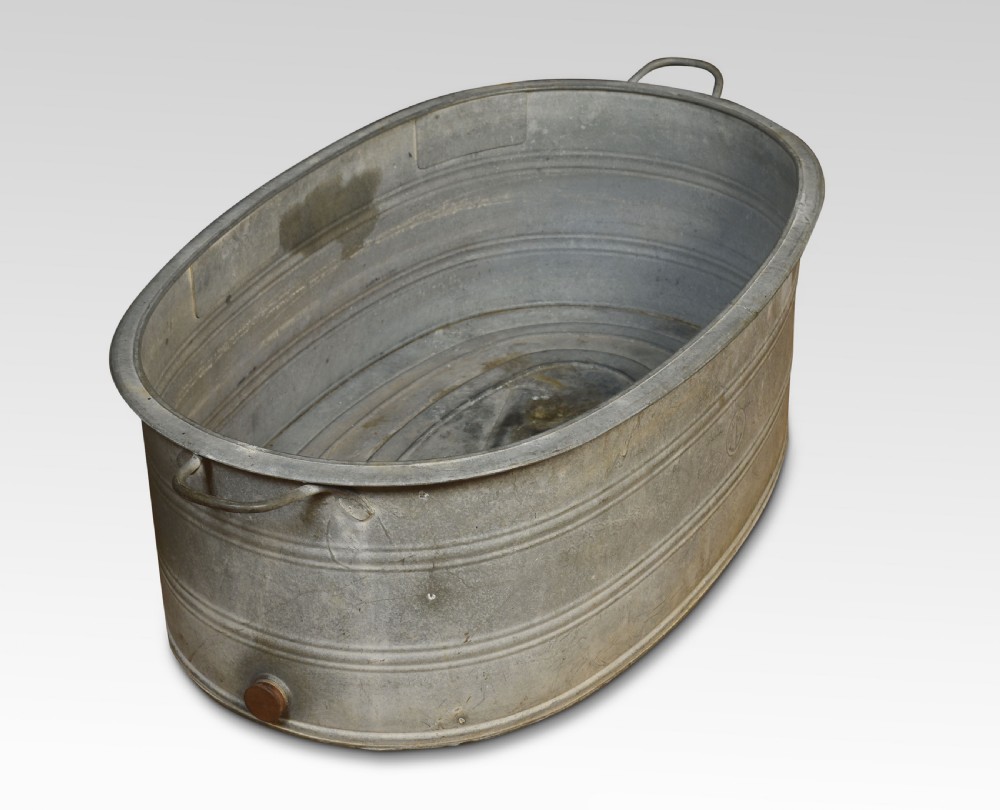 An oval galvanised steel bath tub featuring side carrying handles, ribbed horizontal reinforcement bands, a rolled rim, and a drainage plug near the base. View III