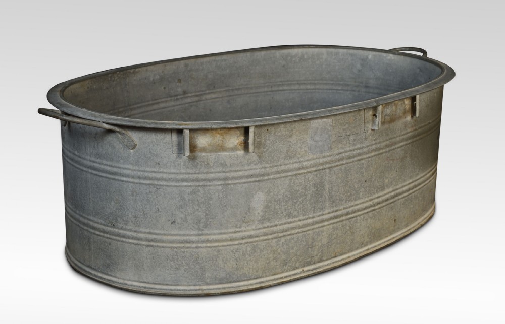 An oval galvanised steel bath tub featuring side carrying handles, ribbed horizontal reinforcement bands, a rolled rim, and a drainage plug near the base. View V