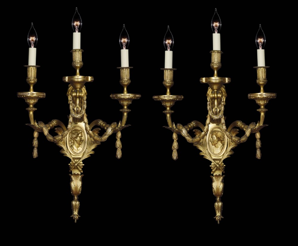 A pair of gilded bronze Louis XVI style three-arm wall lights decorated with classical profile medallions, laurel swags, and acanthus leaf details.