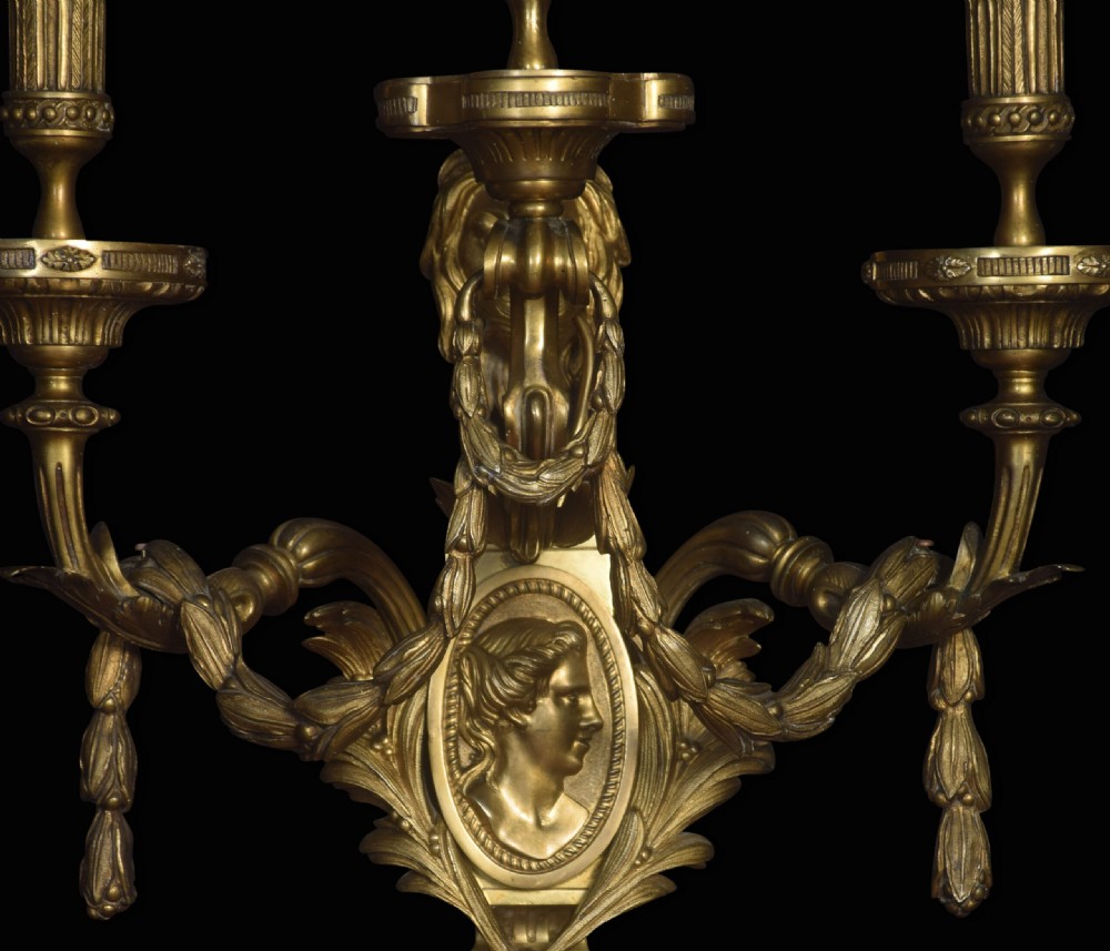 A pair of gilded bronze Louis XVI style three-arm wall lights decorated with classical profile medallions, laurel swags, and acanthus leaf details. View II
