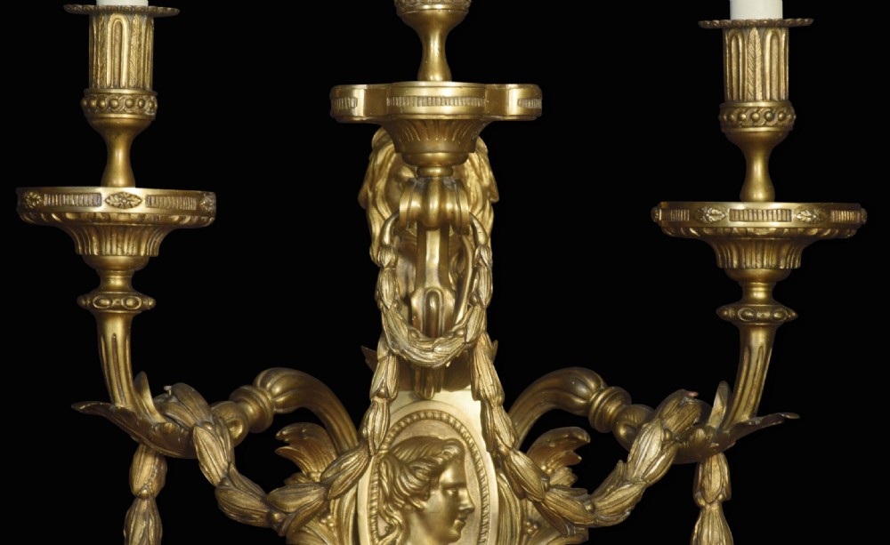 A pair of gilded bronze Louis XVI style three-arm wall lights decorated with classical profile medallions, laurel swags, and acanthus leaf details. View V