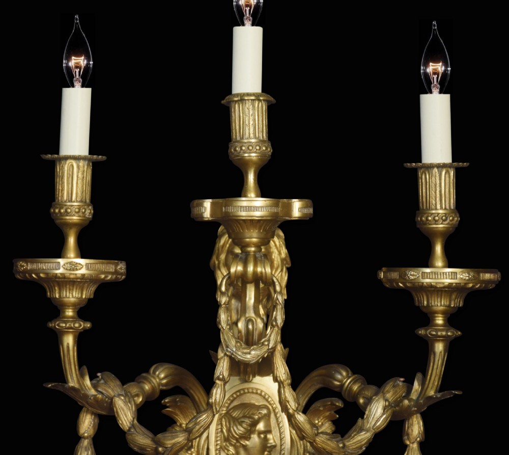 A pair of gilded bronze Louis XVI style three-arm wall lights decorated with classical profile medallions, laurel swags, and acanthus leaf details. View VI
