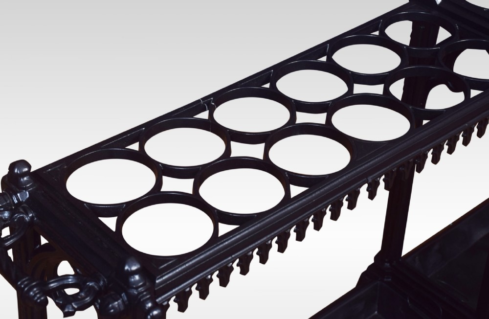 A long black cast iron umbrella stand featuring twenty-four circular rings supported by decorative pierced side panels and integrated base drip trays. View IV